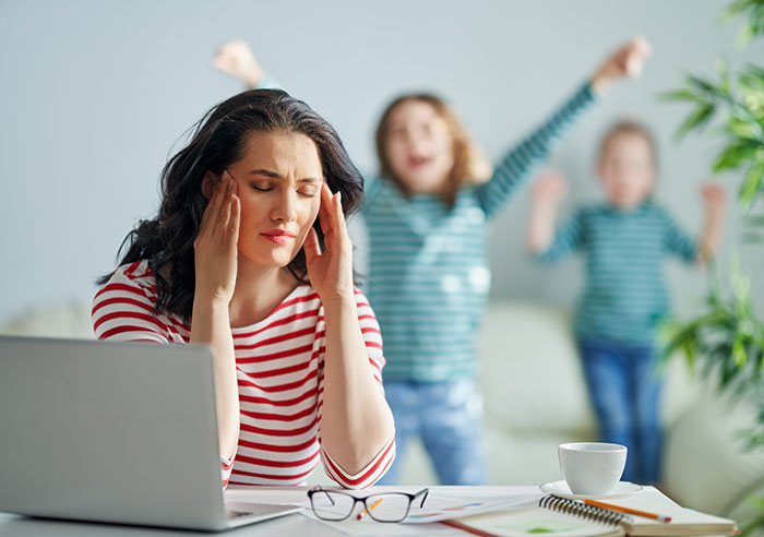 Stressed woman holding her head with kids playing wildly behind her, relating to charging friend for furniture damage.