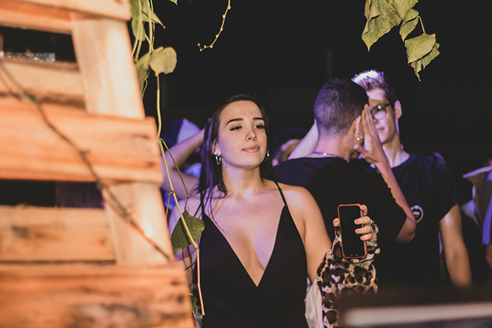 Young woman in a black dress holding a phone, at a nighttime event with people in the background showing mixed emotions.