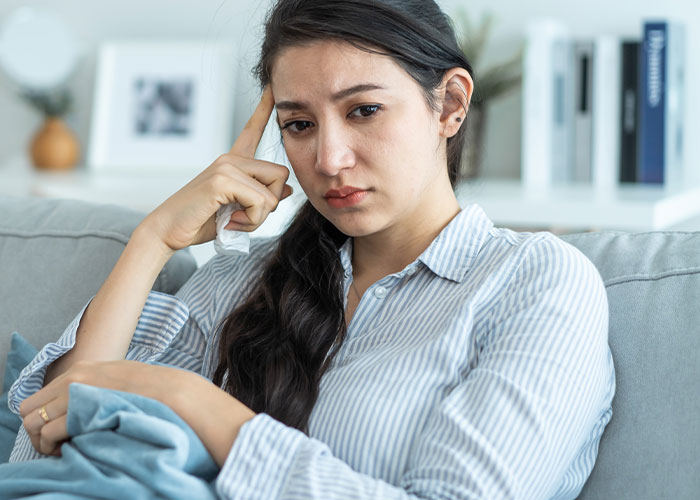 Sad woman sitting on couch looking thoughtful and upset, related to husband&rsquo;s childhood best friend favor humiliation story.