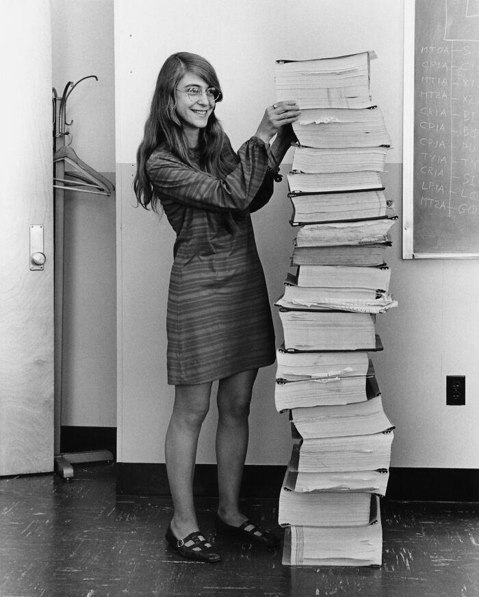 Vintage photo of a woman smiling while stacking a tall pile of large thick books in an office setting.