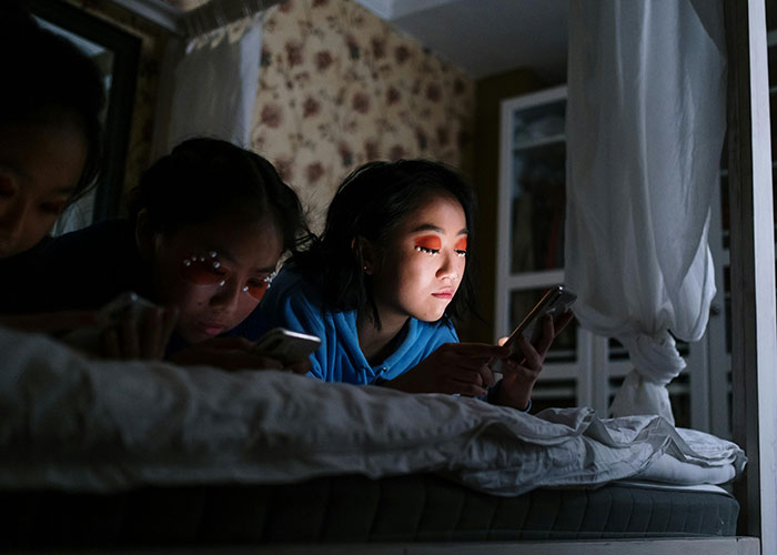 Three young people lying on a bed at night, using smartphones with focused expressions, highlighting Gen X lifestyle contrast.