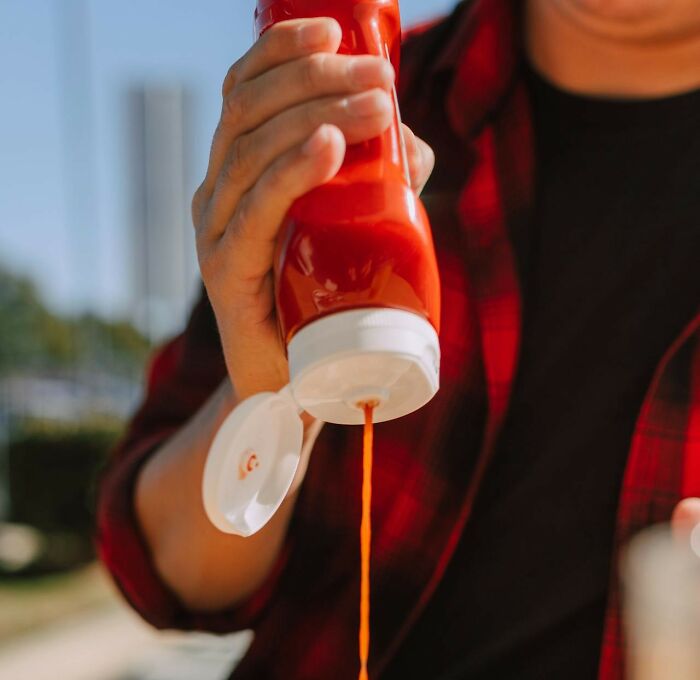 Person in a red shirt squeezing ketchup bottle lazily outside, illustrating excessive laziness in everyday tasks.