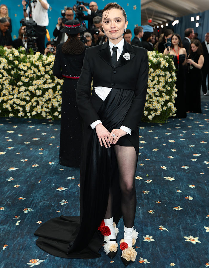 Aimee Lou Wood posing at a formal event wearing a black suit dress with floral shoes on a blue floral carpet. Aimee Lou Wood posing at a formal event wearing a black suit dress with floral shoes on a blue floral carpet.