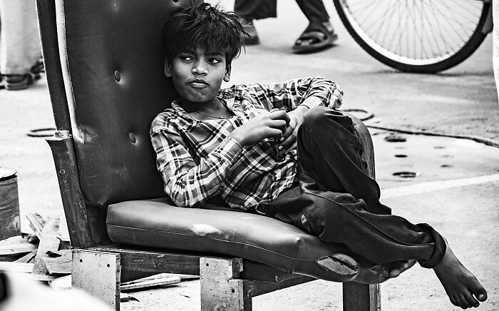 Boy in ripped pants lounging on a worn chair on the street, captured in a striking street photography moment.