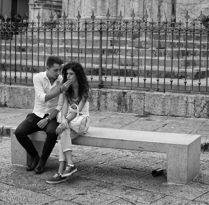 Young couple sharing a moment on a stone bench in a candid street photography scene with historic backdrop.