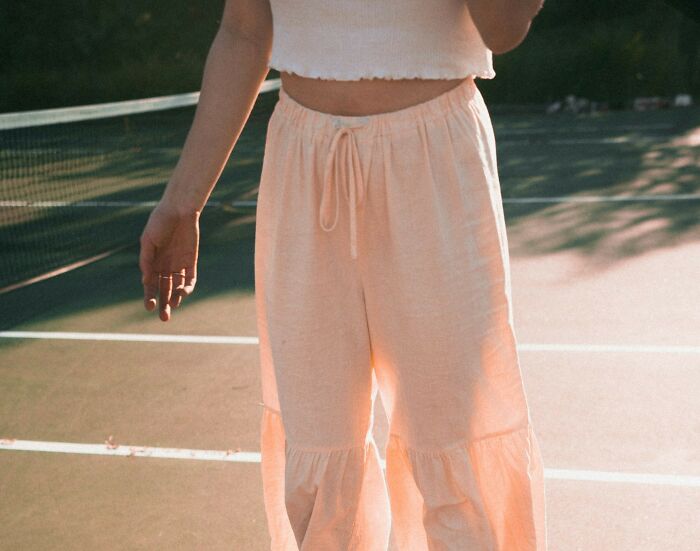 Person wearing light pink pants and cropped top standing on a sunlit tennis court with net in the background, boomer opinions concept.