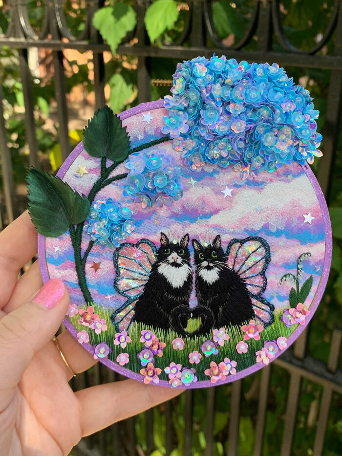 Hand holding embroidery design featuring two black and white cats with fairy wings surrounded by flowers and blue hydrangeas.