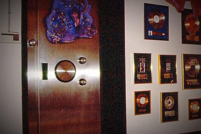 Metal wall with an artistic mask and multiple gold and platinum music records displayed on a white wall nearby.