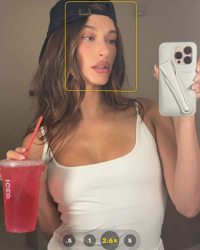 Hailey Bieber taking a bikini selfie wearing a white top and black cap, holding a red iced drink with a straw. Hailey Bieber taking a bikini selfie wearing a white top and black cap, holding a red iced drink with a straw.