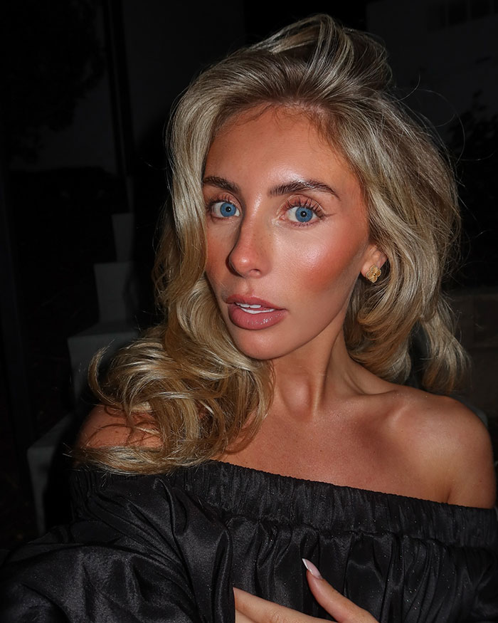 Blonde woman with blue eyes posing in a black off-shoulder top in a dimly lit setting related to Bonnie Blue arrest update.