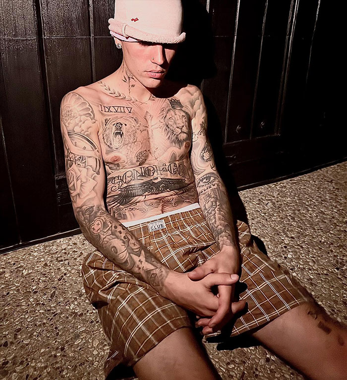 Justin Bieber shirtless in LA, showing tattoos and wearing a pink hat and brown checkered shorts at night.