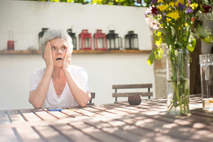 Elderly woman sitting at a wooden table outdoors looking shocked, reflecting the couple goes ballistic wedding dispute.