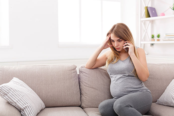 Pregnant young woman sitting on couch looking worried while talking on phone about pregnancy announcement and gender reveal.