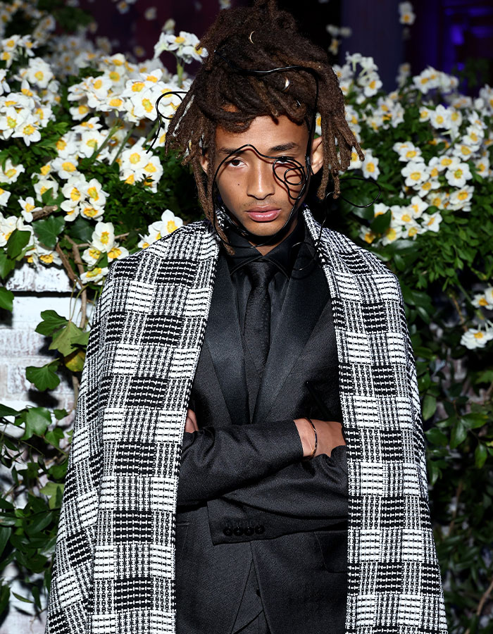 Individual at Met Gala 2025 wearing black suit with checkered coat and artistic face accessory, showcasing fashion fails.