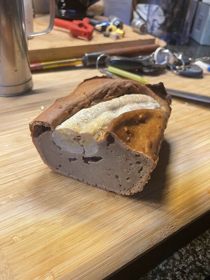 Partially peeled banana baked inside dense, oddly textured bread on a wooden cutting board in a kitchen setting, cursed food pic.