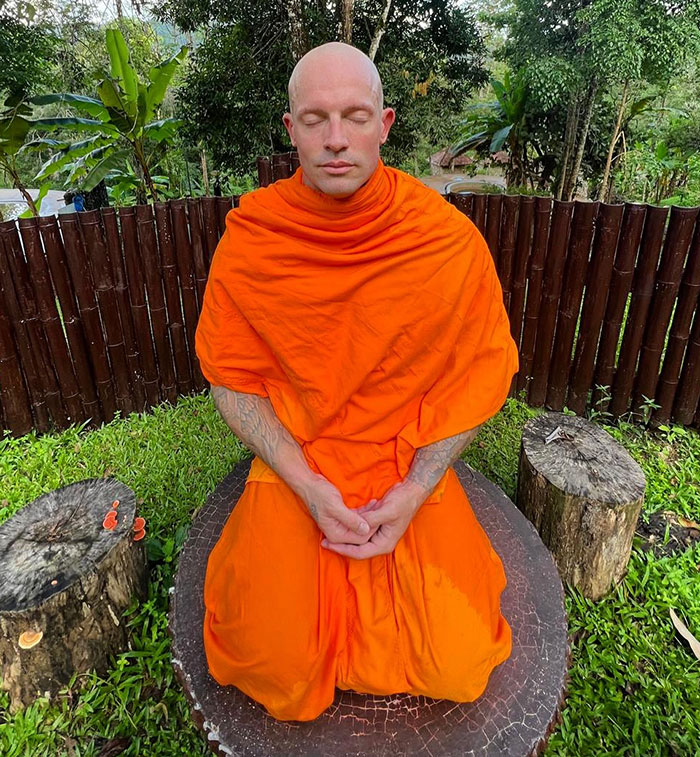 Man formerly a pole dancer now a monk, meditating outdoors in bright orange robes surrounded by nature and wooden logs.