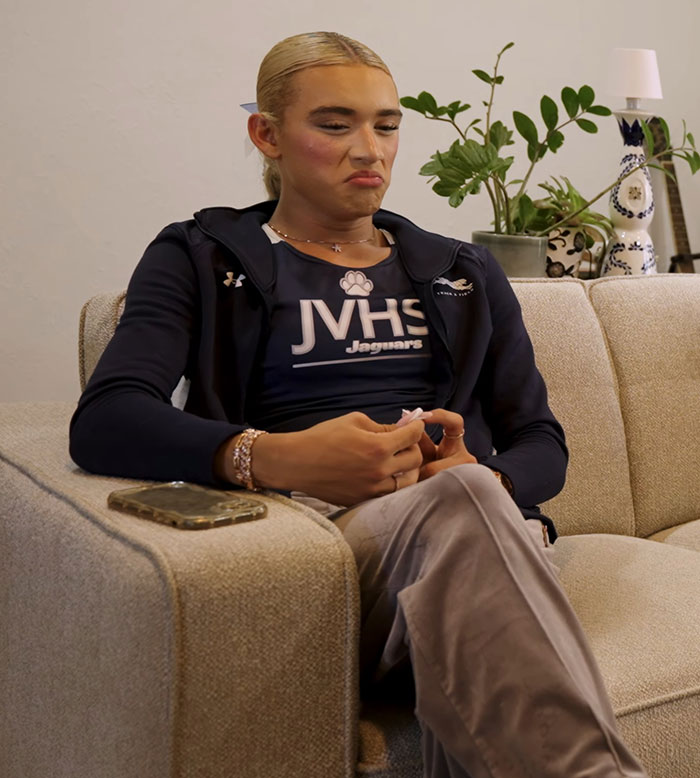 High school girl in JVHS Jaguars gear sitting on couch, showing defiant expression after losing to trans athlete.