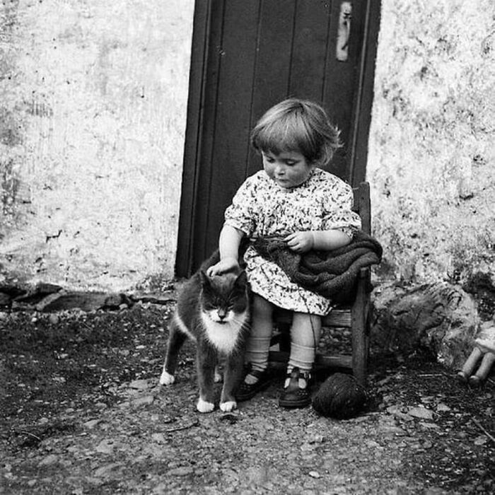 Vintage photo of a young child sitting outdoors, gently petting a cat, showcasing the timeless bond between kids and cats.