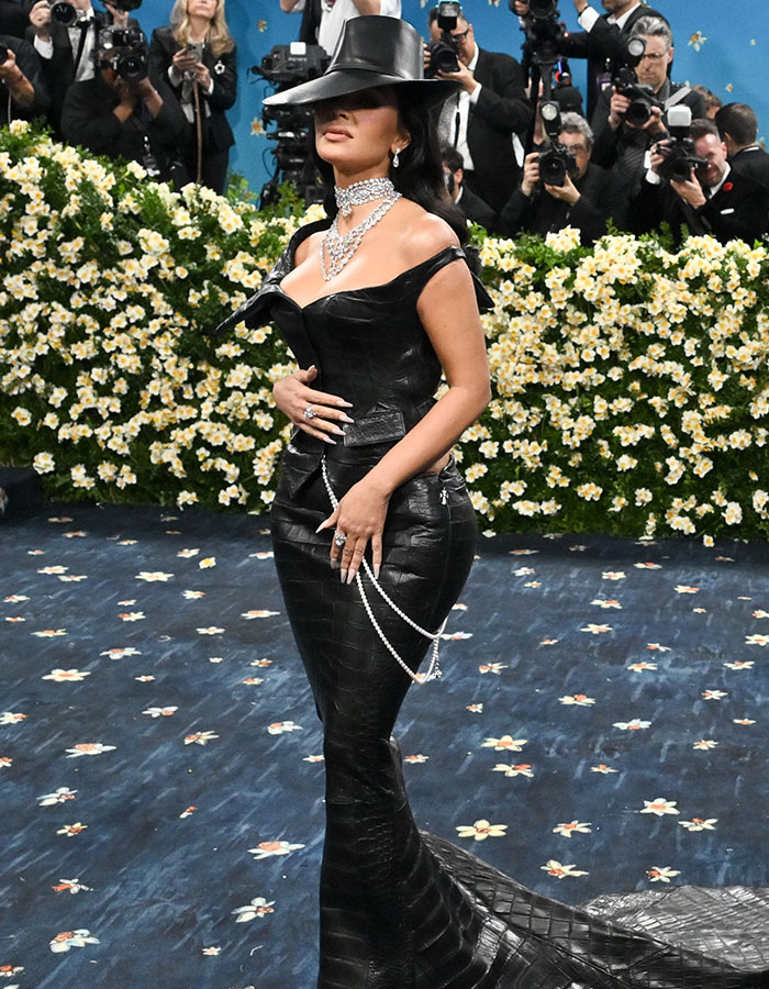 Woman in a black leather gown and hat posing on the Met Gala carpet among photographers, showcasing fashion fails.