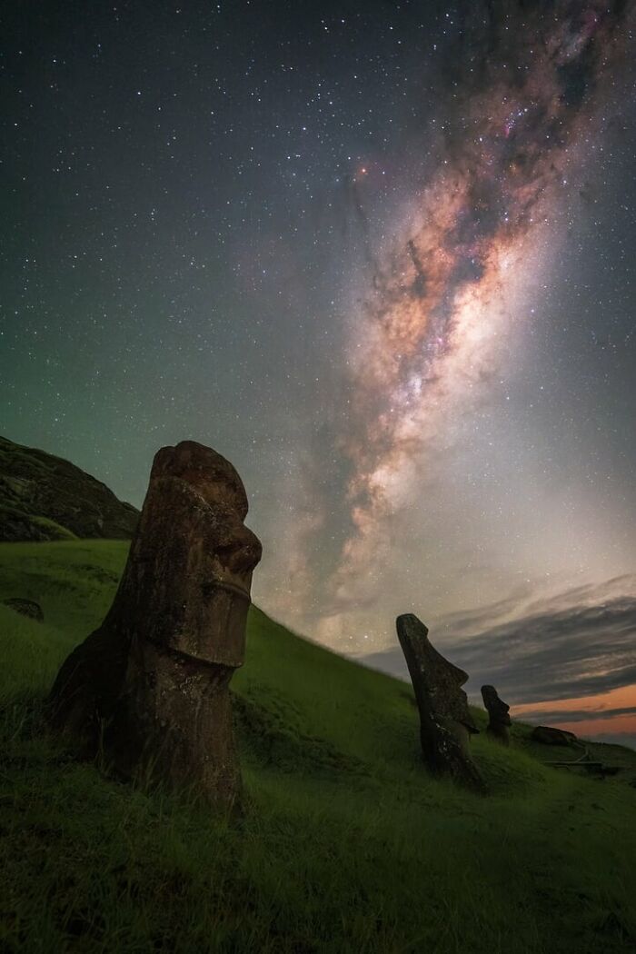 Milky Way glowing above ancient stone Moai statues on grassy hillside under a clear night sky full of stars.