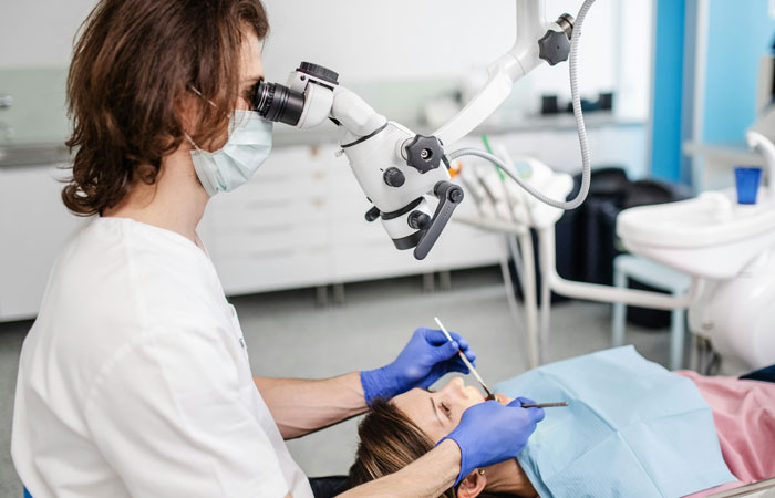 Dentist wearing gloves and mask using microscope while treating a patient, illustrating ways people lose money thinking they’re saving.