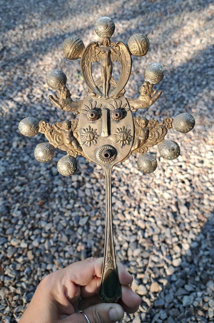 Intricately designed weird and wonderful thrifted find with ornate angel and sun face details held outdoors on gravel.