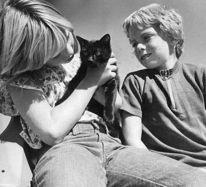 Two kids holding and looking at a black cat in a vintage black and white photo showing timeless bond between kids and cats.