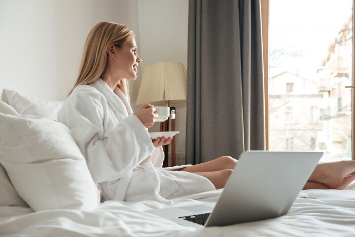 Woman in a white robe enjoying coffee on bed with laptop nearby, a tiny thing that made her trip better and relaxing