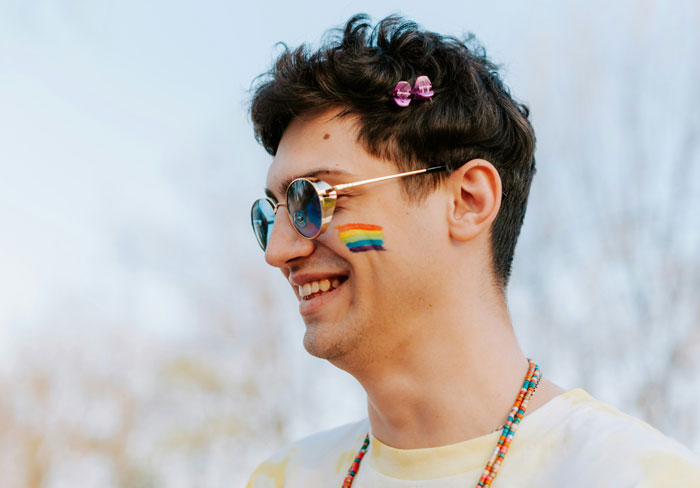 Young man wearing sunglasses with rainbow face paint and beads, smiling outdoors, highlighting problematic behaviors women get a pass for.