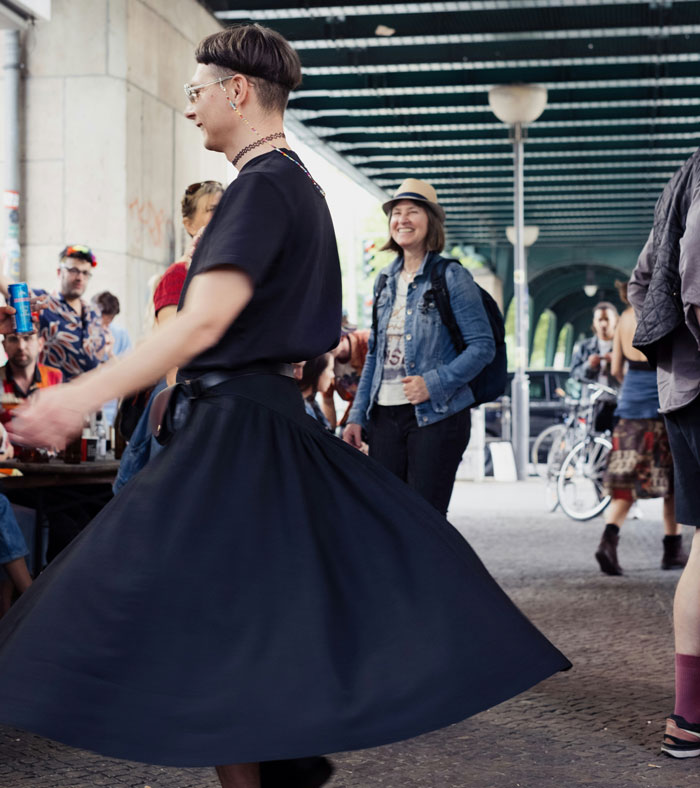 Person spinning in a black skirt under a bridge, with people socializing and bicycles in the background, secret spouse kept.