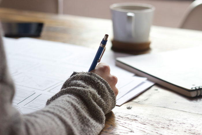 Person in a gray sweater writing with a blue pen on paper at a wooden table with a coffee cup and notebook nearby, illustrating things said by people in charge.