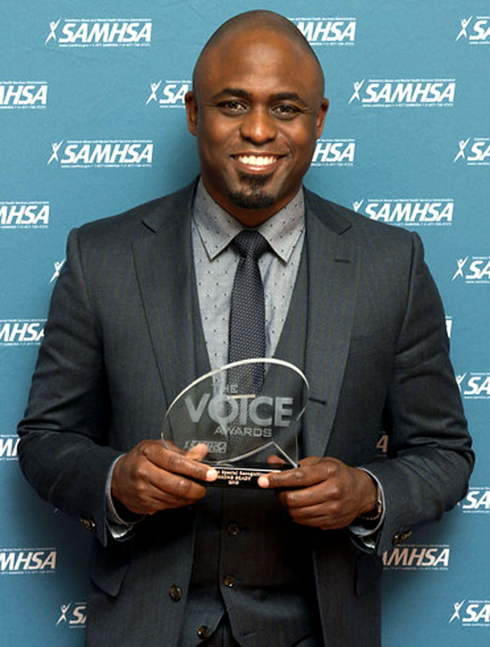 Man in suit holding a Voice Awards trophy at a SAMHSA event, smiling during a positive celebrity encounter moment
