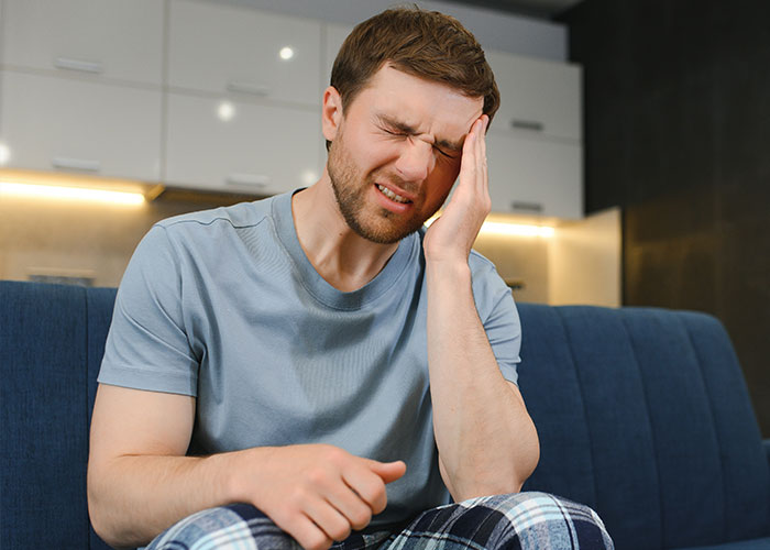 Young man in casual clothes sitting on couch holding his head in pain showing a pathetic injury experience