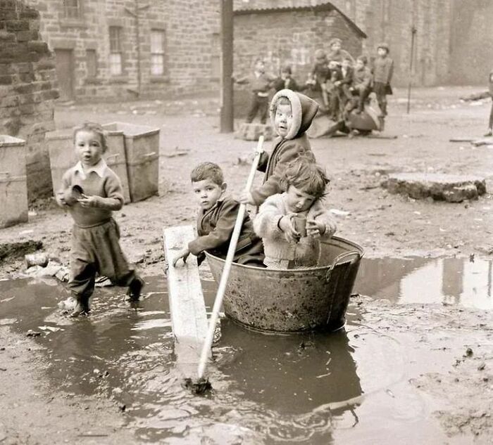 Children playing in a muddy puddle with makeshift boat and paddle, showing a side of history not in textbooks.