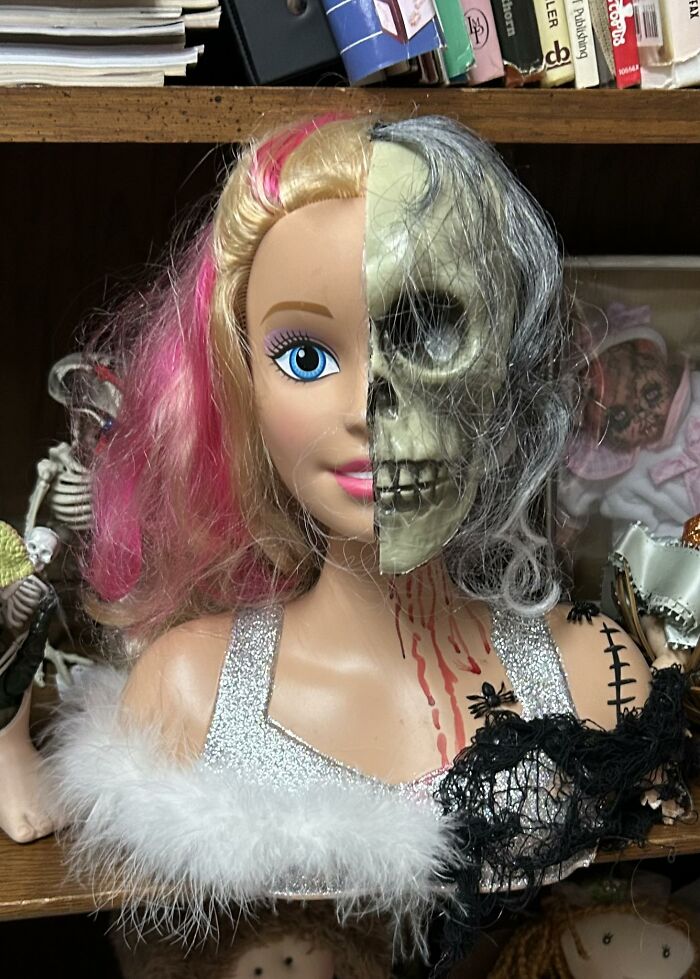 Half doll half skull with pink hair and sparkly silver dress among weird and wonderful thrifted finds on a shelf.