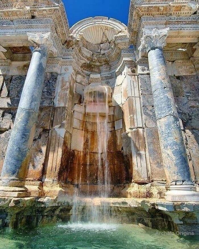 Ancient history ruins with a stone waterfall fountain flowing into a clear blue water basin under bright sunlight.