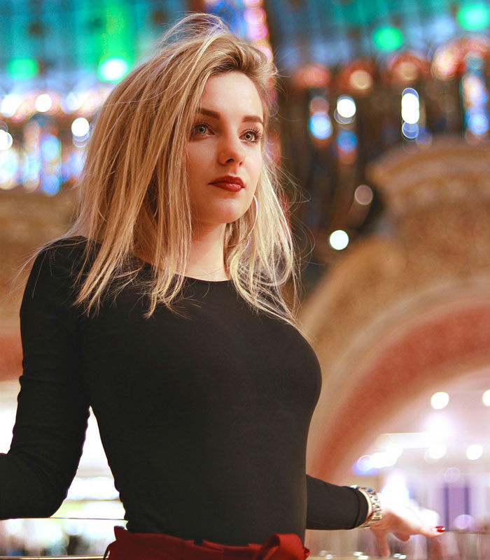 Young woman with blonde hair wearing a black top, posing indoors with colorful lights in the background, depicting problematic behaviors.
