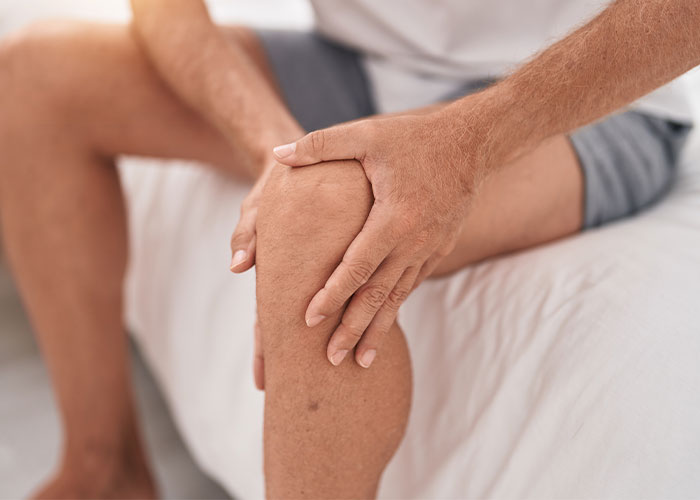 Man sitting on bed holding his knee, illustrating one of the most pathetic injuries people have experienced.