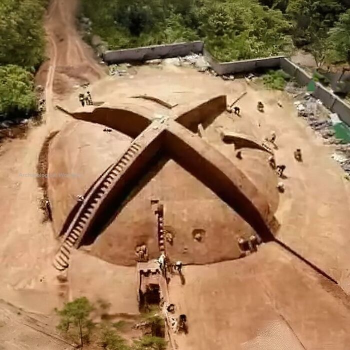 Aerial view of an ancient archaeological site excavation showing a large cross-shaped structure surrounded by trees and pathways.