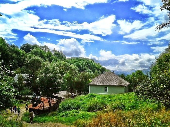 Rural landscape with lush greenery, a traditional house, and visitors walking under a bright blue sky at a UNESCO treasure site.