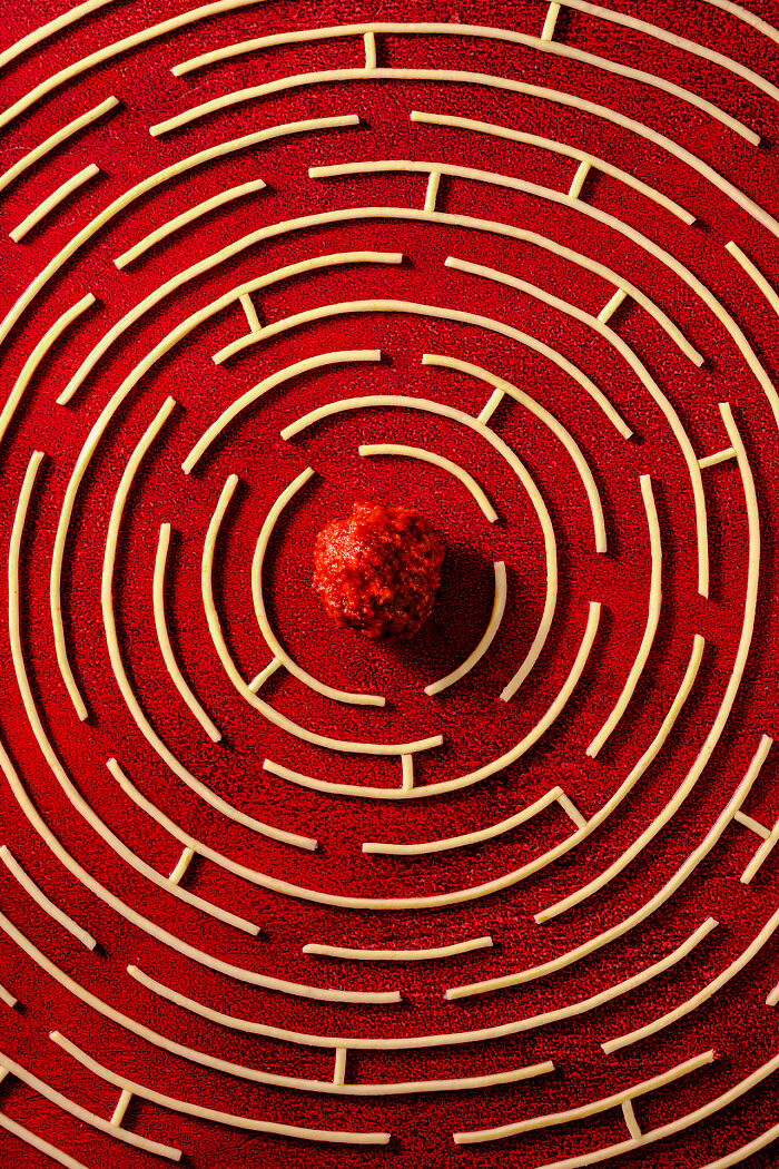 Spaghetti arranged in a circular maze pattern with a meatball at the center, vibrant red background food photography.