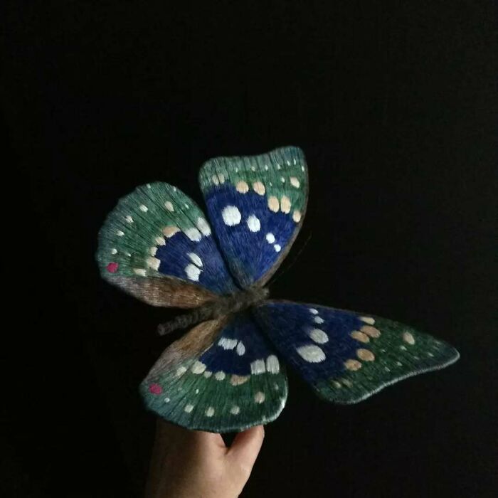 Hand holding a detailed embroidered fiber moth with blue and green wings against a black background.