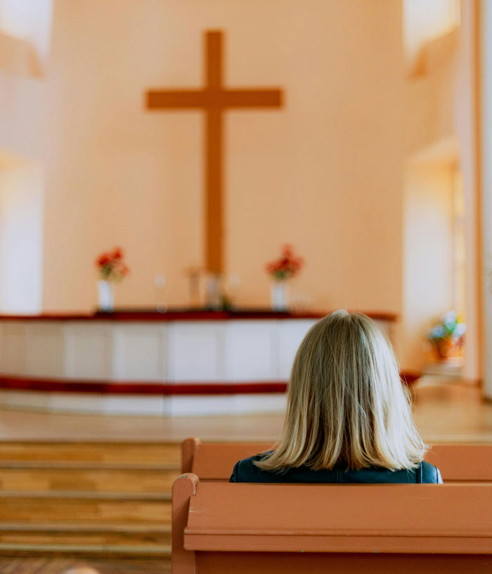 Woman sitting alone in a church pew, facing a large cross, reflecting themes of people who escaped from cults.