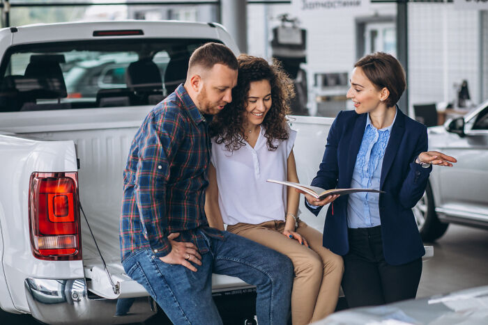 A man showing a white car to a woman inside a showroom, highlighting everyday items that have become expensive.