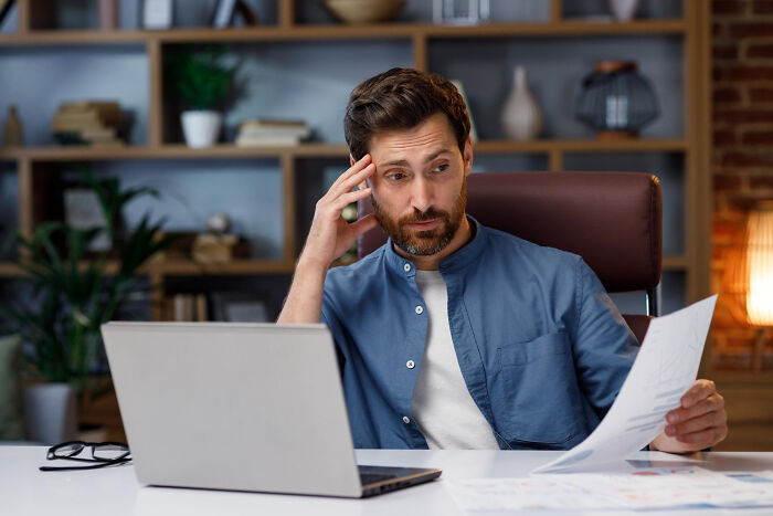 Man in a blue shirt looking stressed while reviewing papers at his desk with laptop, symbolizing life blunders secrets.