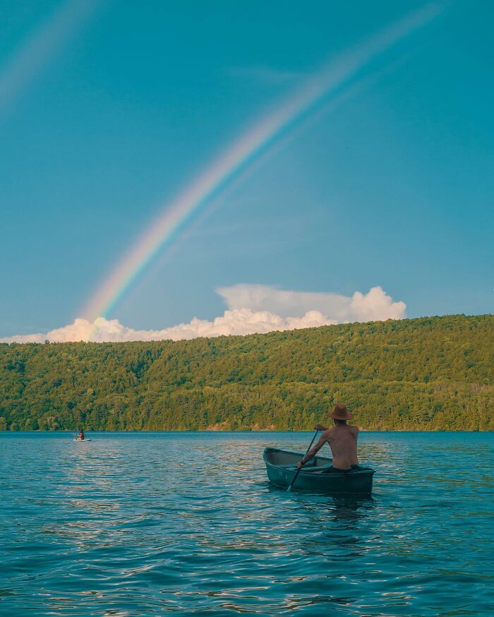 Person paddling a canoe on a lake under a rainbow with a forested hill in the background, creative photo edit.