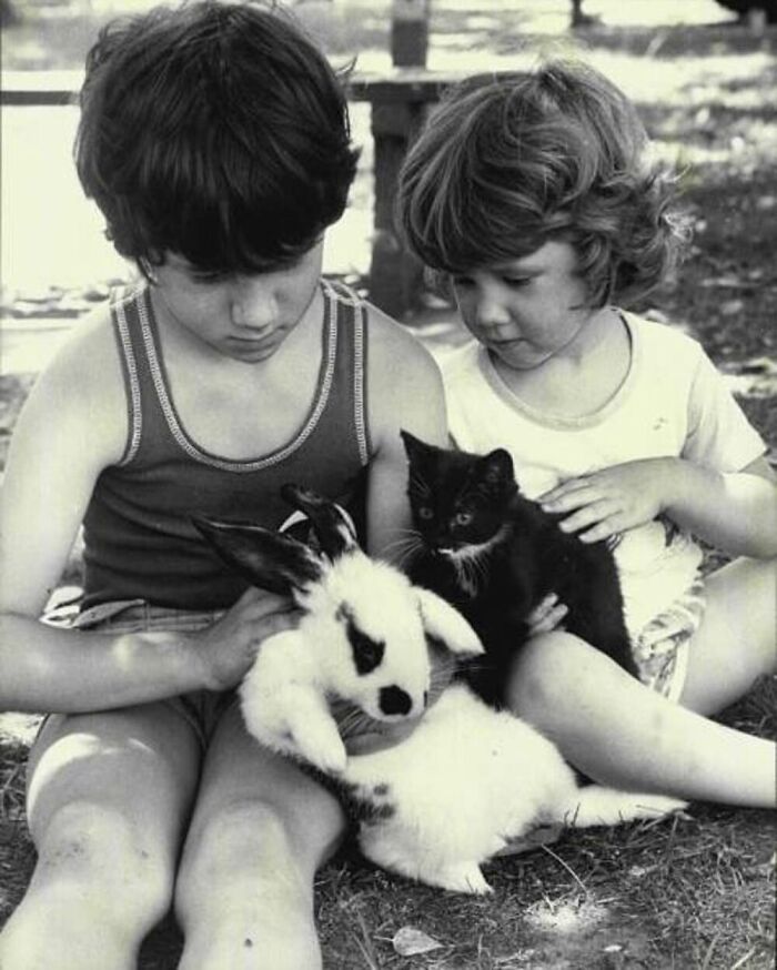 Two kids sitting outdoors holding a black cat and a white rabbit in a vintage photo showing the bond between kids and cats.