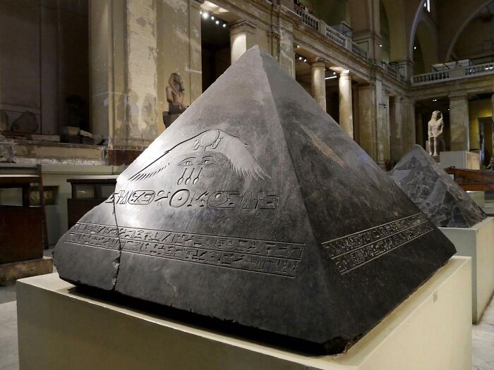 Dark stone pyramid artifact with ancient Egyptian hieroglyphs displayed in a museum of historical artifacts.