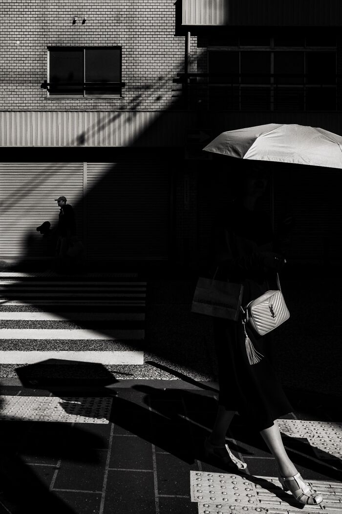 Street photography scene with strong shadows, featuring a woman holding an umbrella and a pedestrian crossing a street.