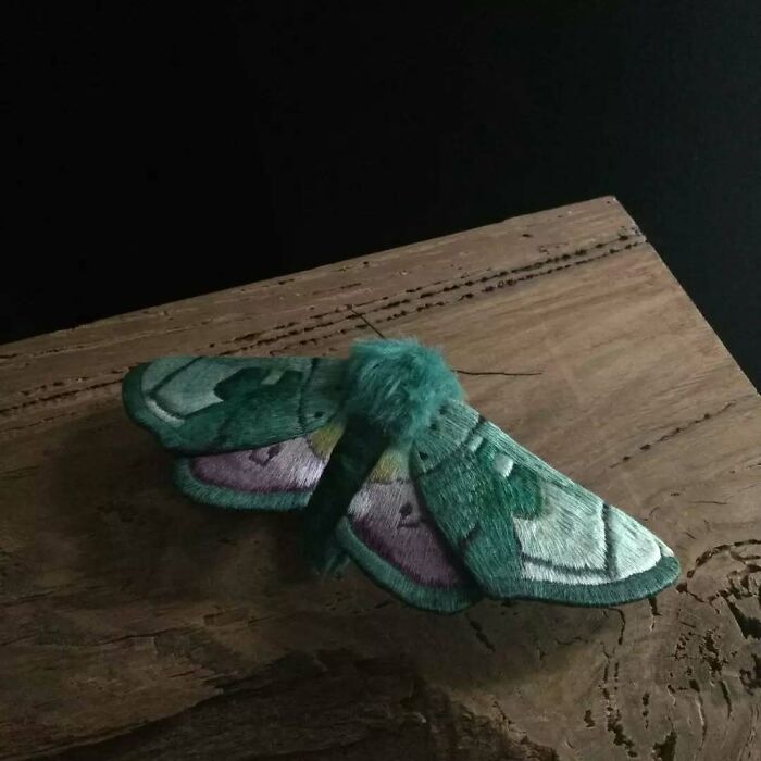 Embroidered fiber moth with green and purple details resting on textured wood, showcasing delicate textile artistry.